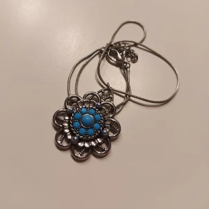 Silverhalsband med turkos blomma - Elegant silverhalsband med ett vackert blomformat hänge. Hänget har turkosa stenar i mitten och en detaljerad design runtom. Perfekt för att ge en färgklick till din outfit. Skriv om ni har någon fundering!✨️