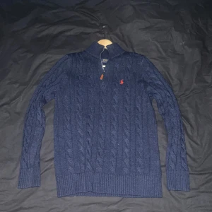 Mörkblå stickad tröja från Ralph Lauren - Säljer en snygg mörkblå stickad tröja från Ralph Lauren med dragkedja och röd broderad logga på bröstet. Tröjan har en ribbad krage och långa ärmar, perfekt för kyligare dagar. Passar både till vardags och mer uppklädda tillfällen.