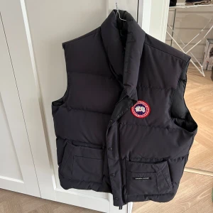 Svart dunväst Canada goose  - Säljer en svart dunväst av märket Canada goose . Västen har en hög krage och praktiska fickor framtill. Perfekt för kyligare dagar.