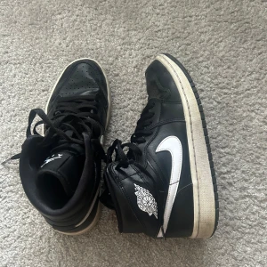 Svarta Nike Air Jordans 1 - Snygga svarta Nike Air Jordans med vit sula och vit swoosh. Skorna har klassisk snörning och perforerade detaljer på tån för extra ventilation. Perfekta för en sportig look.