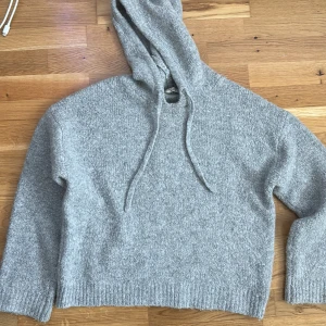 Grå stickad hoodie - Mysig grå stickad hoodie med luva och dragsko. Perfekt för kyliga dagar med sin mjuka och bekväma passform. Ribbstickade muddar vid ärmslut och nederkant ger en extra touch av stil.