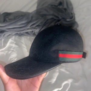 Svart Gucci keps med GG-mönster - Svart keps från Gucci med diskret GG-monogram över hela tyget och klassiska gröna och röda ränder på sidan. Justerbar rem bak i svart skinn med metallspänne. Snygg och stilren accessoar som lyfter din outfit.