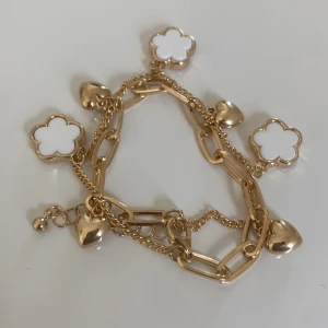 Guldigt armband med blommor och hjärtan - Snyggt armband i guldton med chunky länkar och tunna kedjor. Pryds av vita blommor och blanka hjärtberlocker som ger en lekfull och trendig vibe. Perfekt accessoar för att lyfta din outfit med lite extra bling och sötma.