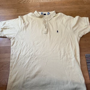 Beige pikétröja från Ralph Lauren - Klassisk beige pikétröja från Ralph Lauren med broderad marinblå logga på bröstet. Tröjan har krage, knäppning med två knappar och korta ärmar. Perfekt för en clean och stilren look.
