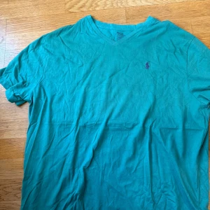 Turkos t-shirt från Polo Ralph Lauren - Turkos t-shirt från Polo Ralph Lauren med klassisk v-ringning och broderad logga på bröstet. Skön bomullskvalitet och normal passform, perfekt till jeans eller shorts. Enkel och stilren design som funkar året runt.