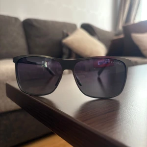 Svarta solglasögon Porsche Design - Snygga svarta solglasögon från Porsche Design med rektangulära bågar och mörka tonade glas. Klassisk och stilren look med diskret logga på skalmen. Lätta och bekväma att bära, perfekta för soliga dagar.