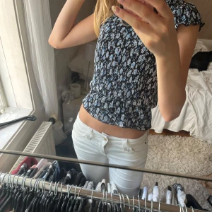 Blommig smockad topp med volangärm - Supersöt smockad topp med blått och vitt blommönster på svart botten. Toppen har korta volangärmar och en lätt croppad passform. Perfekt för dig som gillar feminina detaljer och mönster. Sitter tajt men stretchigt över kroppen. Aldrig använd!❣️