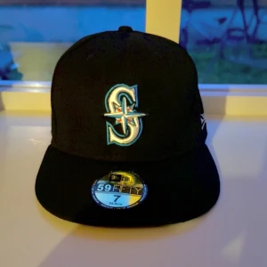 Svart New Era Seattle Mariners keps - Snygg svart 59FIFTY keps från New Era med Seattle Mariners-logga broderad framtill i vitt, turkost och silver. Klassisk flat peak och MLB-logga baktill. Perfekt för dig som gillar baseball eller vill ha en stilren streetwear-accessoar. Helt ny 