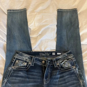 Miss Me jeans med broderade bakfickor - Snygga blå jeans från Miss Me med vita kontrastsömmar och stora broderade detaljer på bakfickorna. Jeansen har raka ben och coola nitar på fickorna. Perfekta för dig som vill sticka ut med din denimstil.