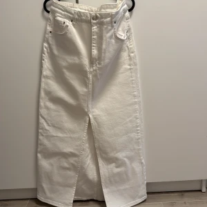 Vit jeanskjol från Gina Tricot - Snygg vit lång jeanskjol från Gina Tricot med klassiska fickor fram och bak, hög midja och slits framtill. Perfekt för dig som gillar stilren och trendig denim-look. Kjolen är i kraftig bomull och har knapp- och dragkedjestängning.