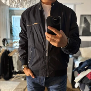 Svart bomberjacka från Vailent Purex - Snygg svart bomberjacka från Vailent Purex med silvriga dragkedjor och ribbade muddar. Jackan har en minimalistisk look, ståkrage med tryckknappar och meshfoder på insidan. Perfekt för dig som gillar stilren och urban vibe.