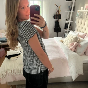 Svartvit rutig blus med v-ringning - Supersöt blus i svartvitt rutigt mönster med korta ärmar och v-ringad hals. Lätt och luftig modell som faller snyggt över kroppen. Perfekt till jeans eller kjol för en chill och trendig look. Från Bondelid. 