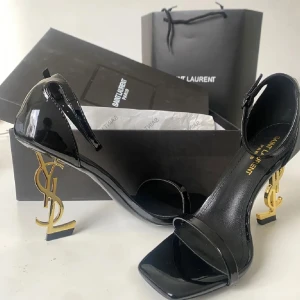 Svarta Saint Laurent sandaler med YSL-klack - Snygga svarta Saint Laurent-sandaler i lackat skinn med öppen tå och fyrkantig front. Klacken är 8 cm hög, i guld och formad som YSL-loggan för en lyxig look. Smal rem över tårna och justerbar ankelrem för bra passform.