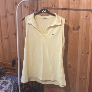 Gul ärmlös pikétopp från Lacoste - Snygg gul ärmlös pikétopp från Lacoste med klassisk krage och knappar framtill. Toppen har det ikoniska krokodilmärket broderat på bröstet och är gjord i bomull. Perfekt för en sportig och fräsch look under varma dagar.