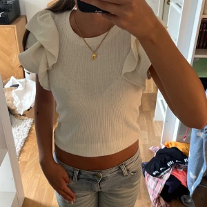 Vit ribbad topp med volangärmar - Supersöt vit ribbad topp med korta volangärmar som ger en extra touch till outfiten. Modellen är croppad och har rund halsringning. Perfekt för dig som gillar en feminin och trendig stil.