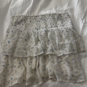 Blommig volangkjol från Vero Moda - Supersöt kort kjol från Vero Moda med volanger och små blå blommor på vit botten. Kjolen har smock i midjan för en bekväm passform och är perfekt för varma dagar. Lätt och luftig känsla, passar perfekt till sneakers eller sandaler.