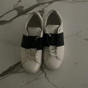 Valentino Garavani Open sneakers vit/svart - Snygga Valentino Garavani Open sneakers i vitt skinn med bred svart läderdetalj över snörningen. Platta sulor, rund tå och ikoniska nitar baktill ger en lyxig och modern vibe. Perfekta för dig som vill sticka ut med stilrena och exklusiva sneakers.