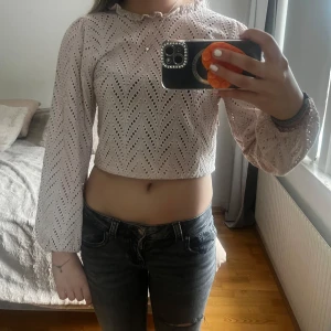 Ljusrosa croppad blus med volang - Säljer en ljusrosa/beige croppad blus med långärmade puffärmar och broderat hålmönster. Toppen har volangdetaljer vid halsringningen och en öppen rygg som ger en extra snygg touch. Perfekt för dig som gillar romantisk och trendig stil.