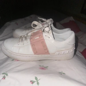 Valentino Rockstud sneakers vit/rosa - Snygga Valentino Rockstud sneakers i vitt skinn med rosa band och ikoniska nitar. Skorna har rund tå, snörning och platt sula. Perfekta för dig som vill ha en lyxig och trendig look med en touch av rosa detaljer.