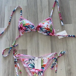 Blommig bikini från Love by Lola - Superfin bikini med tropiskt blommigt mönster i rosa, grönt, blått och orange. Triangeltopp med knyt i nacke och rygg samt matchande trosa med knyt i sidorna. Söt rosa spetskant runt både topp och trosa. Perfekt för sommaren, storlek XL men sitter även bra på en M eftersom man själv knyter banden!💗(orginal pris 1000kr)