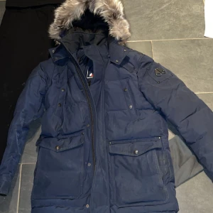 Blå Moose Knuckles dunjacka med pälskant - Säljer en mörkblå Moose Knuckles med fet pälskantad huva i storlek M, jackan är bara använd en vinter, säljer för jag har gått ner i vikt och passar inte mig längre lika bra