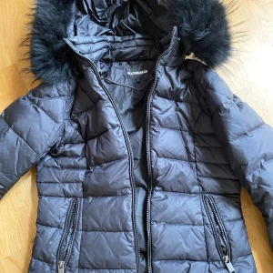 Svart dunjacka Rockandblue med fuskpäls - Snygg svart dunjacka från Rockandblue i storlek 36. Jackan har quiltad design, dragkedjor framtill och på fickorna samt en huva med fluffig svart fuskpäls. Perfekt för kalla dagar och riktigt stilren look.