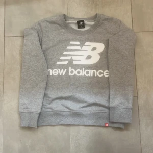 Grå sweatshirt från New Balance XS - Snygg grå sweatshirt från New Balance i storlek XS. Tröjan har stort vitt logotryck framtill, ribbade muddar och rund hals. Perfekt för en Stockholmsstil och avslappnad stil. Tillverkad i mjuk bomullsblandning för extra komfort.