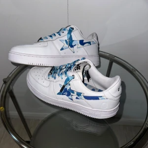 BAPE STA vita sneakers med blå camo - BAPE STA sneakers i vitt skinn med blå och ljusblå camo-detaljer på sidan och matchande blå camo-skosnören. Klassisk låg modell med perforerad tå och tydlig BAPE-logga på plösen och hälen. Perfekta för dig som vill sticka ut med streetwear.