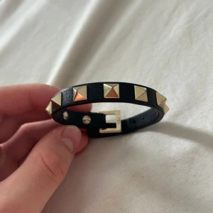 Valentino armband med nitar - Snyggt svart armband från Valentino i äkta skinn med guldiga pyramidformade nitar runt om. Spännet är också i guldton och har en diskret logga. Perfekt accessoar för att ge din outfit en edgy vibe.