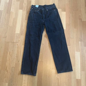 Levi's 565 svart jeans straight fit - Klassiska Levi's 565 jeans i svart denim med raka ben och normal passform. Byxorna har fem fickor, knappgylf och den ikoniska läderpatchen bak i midjan. Perfekta för dig som gillar tidlös stil och vill ha ett par jeans som funkar till allt. Storlek 30/32