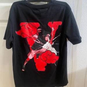 VLONE T-shirt - Svart t-shirt från VLONE med Naruto-inspirerat tryck i rött och vitt. Stor V-logga och animekaraktär på ryggen, samt öga och röda moln på bröstet. Klassisk passform och rund hals. Perfekt för dig som gillar streetwear och anime. Tags och påse medföljer!
