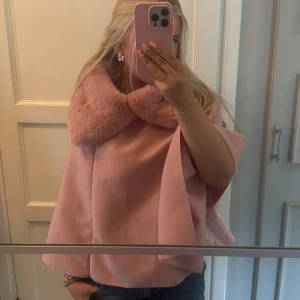 Rosa kappa/poncho med pälskrage - Säljer en superfin ljusrosa kappa/poncho med bred, fluffig pälskrage. Jackan har en loose passform och vida ärmar, vilket ger en trendig och elegant look. Perfekt för dig som vill sticka ut med en mjuk och lyxig vibe.