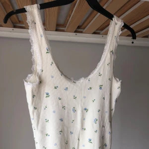 Vit blommig linnetopp från Monki - Supersöt vit linnetopp från Monki med små blå blommor och gröna blad. Toppen har ribbad struktur, spetskant runt halsringning och axelband.