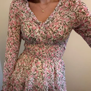 Blommig kortklänning med volanger - Supersöt kortklänning med rosa och grönt blommönster på vit botten. Klänningen har långa ärmar, smockad midja och volangig kjol. V-ringad med knappar framtill och lätt, luftig känsla. Perfekt för sommarens alla tillfällen!