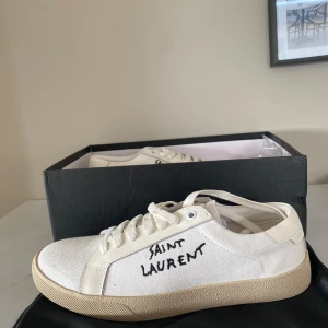 Saint Laurent vita sneakers strl 40 - Snygga vita sneakers från Saint Laurent med handskriven logga på sidan. Skorna har en beige platt sula, snörning och är gjorda i canvas med detaljer i skinn. Perfekta för dig som gillar stilren och trendig design.