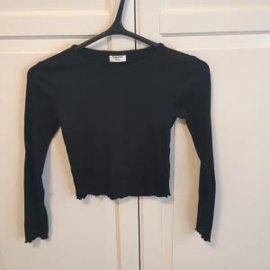 Svart croppad långärmad topp Kappahl - Svart långärmad topp från Kappahl i croppad modell med vågig kant nertill och vid ärmslut. Toppens material är ribbad bomull som ger en skön och stretchig känsla. Perfekt till jeans eller kjol💞
