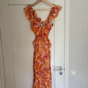 Färgglad långklänning från Zara S - Snygg långklänning från Zara i storlek S med volangdetaljer, knytning fram och öppen rygg. Klänningen har ett färgstarkt mönster i orange, rosa och gult på ljus botten. Perfekt för sommarens alla tillfällen!
