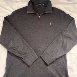 Grå half zip tröja Polo Ralph Lauren - Mörkgrå långärmad tröja från Polo Ralph Lauren med klassisk broderad logga på bröstet. Tröjan har en snygg half zip-dragkedja och ribbade muddar. Perfekt för lager-på-lager och chill stil. Materialet är mjukt och känns riktigt skönt mot huden.