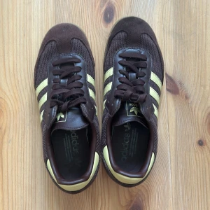 Adidas Samba brun/gul sneakers - Säljer ett par Adidas Samba sneakers i mörkbrunt med ljusgula ränder och detaljer. Ovandelen är i mocka och textil, med klassisk gummisula och snörning. Ikonisk retrostil som passar perfekt till streetwear och vardagsoutfits.