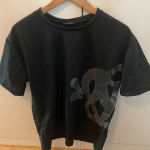 Svart t-shirt med grått tryck, M - Cool svart t-shirt i medium med stort grått grafiskt tryck på både fram- och baksidan. Klassisk rund hals och korta ärmar. Perfekt för dig som gillar streetstyle och vill sticka ut med en unik design.