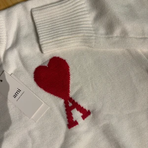 Vit stickad tröja från Ami Paris - Vit stickad tröja från Ami Paris med rund halsringning och ribbade muddar. På bröstet finns ett rött hjärta med bokstaven A, vilket ger en unik och trendig look. Perfekt för dig som gillar stilrena plagg med en twist.