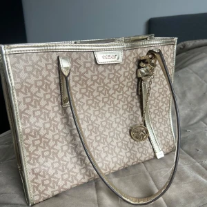 Vintage bag from DKNY - Snygg axelväska från DKNY i beige med guldiga detaljer och logomönster över hela väskan. Väskan har rejäla handtag, dekorativt DKNY-hänge och insida med flera fack. Perfekt för dig som vill ha en stilren och lyxig look.