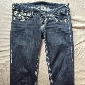 True Religion straight jeans blå, stl 25 - Snygga blå jeans från True Religion med raka ben och tydliga vita kontrastsömmar. Klassiska fickor med lock och silverknappar bak, dragkedja framtill. Ikonisk logga på bakfickan och coola detaljer som sticker ut.