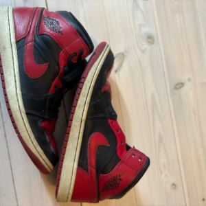 Nike Air Jordan 1 Bred Highs 42 - Säljer ett par klassiska Nike Air Jordan 1 Highs i svart och rött skinn. Ikonisk vinge-logga på sidan och röd swoosh. Hög modell med snörning och vintage-inspirerad sula. Perfekta för dig som vill sticka ut med en tidlös sneaker.