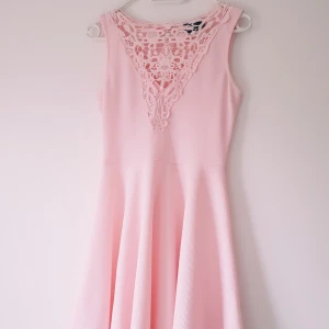 Ljusrosa klänning från AX Paris strl M - Supersöt ljusrosa kortklänning från AX Paris med ärmlös design och vacker spetsdetalj framtill. Klänningen har en lätt utställd kjol och struktur i tyget, samt dragkedja i ryggen. Perfekt för dig som vill ha en feminin och romantisk look.  Trådsläpp finns (se extra bilder) inga hål! Annars fint skick