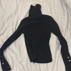 Svart ribbad polotröja med knappar - Svart ribbad polotröja med lång ärm och coola silvriga knappar vid ärmsluten. Tröjan har en hög polokrage och är figurnära i modellen, perfekt för lager på lager. Materialet är stretchigt och mjukt, vilket gör den riktigt skön att bära.