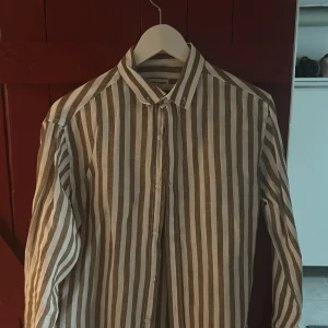 Randig linneskjorta från Bruun & Stengade - Säljer en snygg skjorta från Bruun & Stengade i beige och vitt med breda vertikala ränder. Skjortan är i luftig linne, har klassisk krage, knäppning framtill och långa ärmar. Perfekt för dig som gillar stilren och avslappnad look.