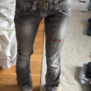 Grå slitna bootcut jeans - Säljer ett par grå jeans med slitningar och hål på knäna. Byxorna har bootcut-modell, låg midja och coola detaljer som dubbla knappar i midjan. Dem är uttsydda på innersidan, vid benen! Hör av er vid frågor elller intresse och kolla gärna in resten av det jag säljer!