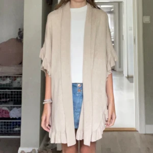 Beige stickad väst med volang från Joseph A. - Beige stickad väst från Joseph A. med snygga volangdetaljer längs kanterna och en knapp för armarna om går att ta ut. Västen är loose fit och har öppen front, perfekt att slänga över en t-shirt eller blus. Materialet är mjukt och skönt, en mix av rayon och nylon.