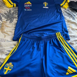 Sverige fotbollströja och shorts Adidas - Säljer ett set med blå och gula Sverige fotbollströja och matchande shorts från Adidas. Tröjan har korta ärmar, tryckta sponsorer och landslagets emblem. Shortsen har gula ränder och Sverige-logga. Materialet är lätt och andas, perfekt för träning eller match.